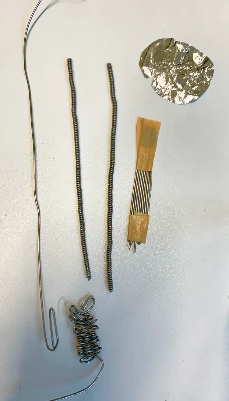 Thin metal wires and coils next to foil and a strip of needles on a white surface