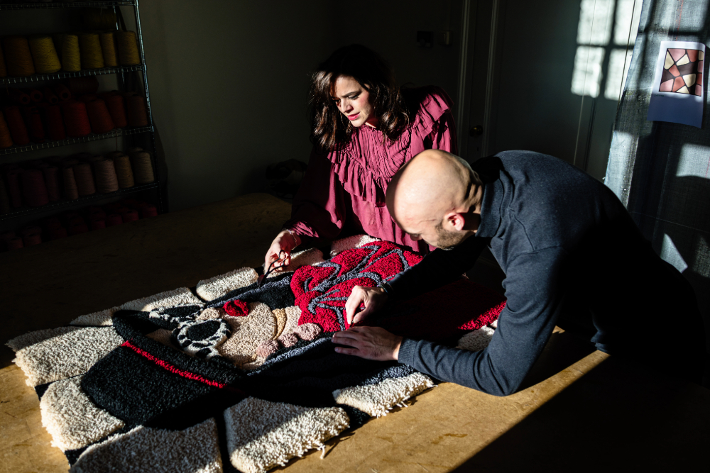 A collaborative moment between two people working on a design. A woman in a blue outfit holds a cup and smiles while seated at a table, while a man in a black shirt leans over, pointing at a paper. On the table are samples of textured materials and spools of yarn in white, beige, and black, reflecting a creative workspace.