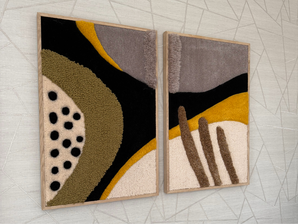 A vibrant textile artwork featuring bold abstract shapes in orange, blue, green, yellow, and purple on a black background. The piece is framed in wood and held under the arm of a person dressed in a dark jacket, with a green ivy wall in the background.
