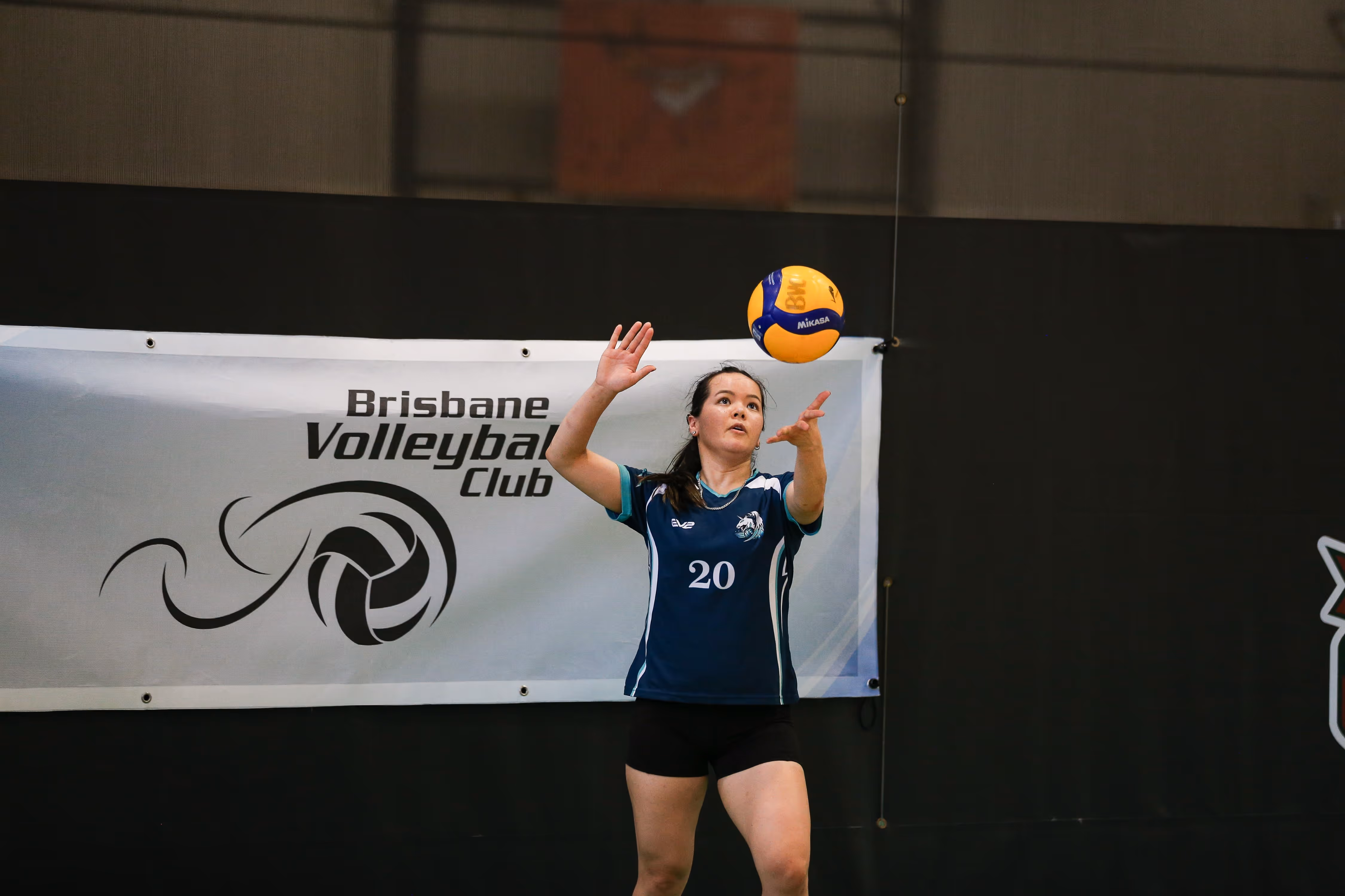 A young female volleyball player serves a ball during a match in front of a Brisbane Volleyball Club banner