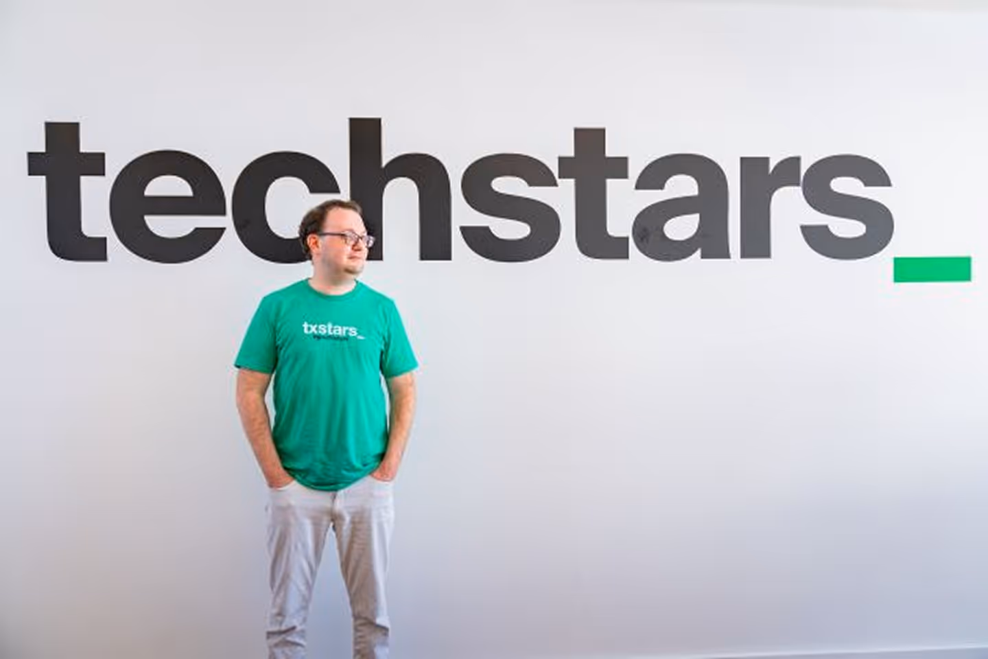 Man wearing green txstars t-shirt standing with hands in pockets in front of a wall with large techstars logo.