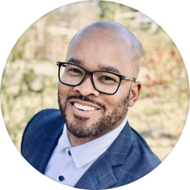 Smiling man with glasses and a beard wearing a blue suit jacket and white shirt outdoors.