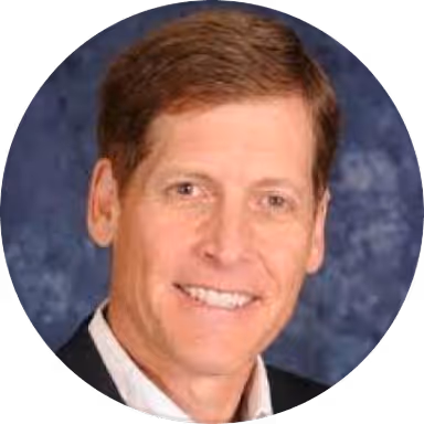 Headshot of smiling middle-aged man with short brown hair wearing a suit jacket and white shirt against a blue background.