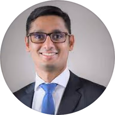 Man wearing glasses, a white shirt, blue tie, and dark suit jacket, smiling against a gray background.