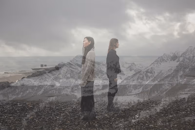 Double exposure of two women standing back to back on a rocky beach with a cloudy sky and a mountain landscape overlay.
