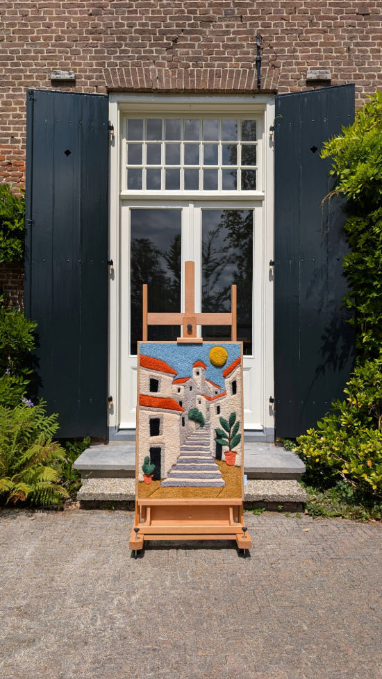 A textile portrait of a woman with a patterned scarf and closed eyes, set on a vivid blue background. The piece is displayed outdoors on a wooden easel against a brick wall with black shutters and white-framed windows