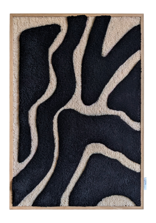 A textile artwork depicting a stylized portrait of a man with a beard, mustache, and large round glasses, wearing a green collared shirt. The piece is displayed outdoors on a wooden easel, set against a brick facade with white-framed glass doors and black shutters, surrounded by greenery.