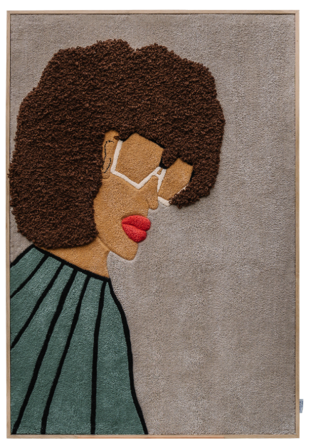 A textile portrait of a woman with a patterned scarf and closed eyes, set on a vivid blue background. The piece is displayed outdoors on a wooden easel against a brick wall with black shutters and white-framed windows