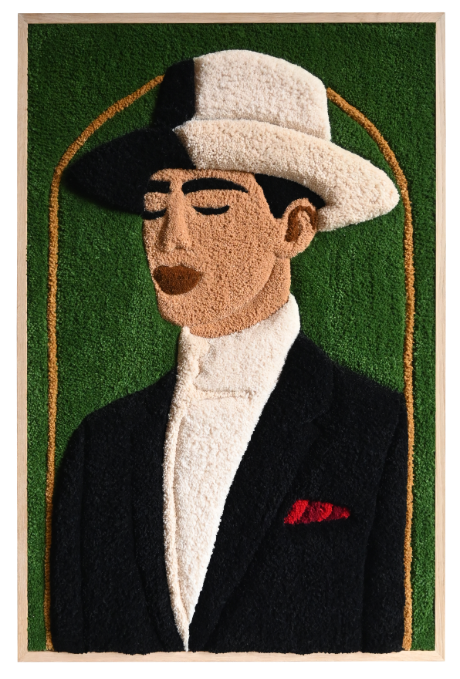 A textile artwork depicting a stylized portrait of a man with a beard, mustache, and large round glasses, wearing a green collared shirt. The piece is displayed outdoors on a wooden easel, set against a brick facade with white-framed glass doors and black shutters, surrounded by greenery.