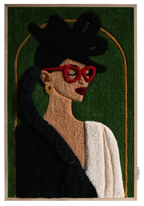 A textile portrait of a woman with a patterned scarf and closed eyes, set on a vivid blue background. The piece is displayed outdoors on a wooden easel against a brick wall with black shutters and white-framed windows