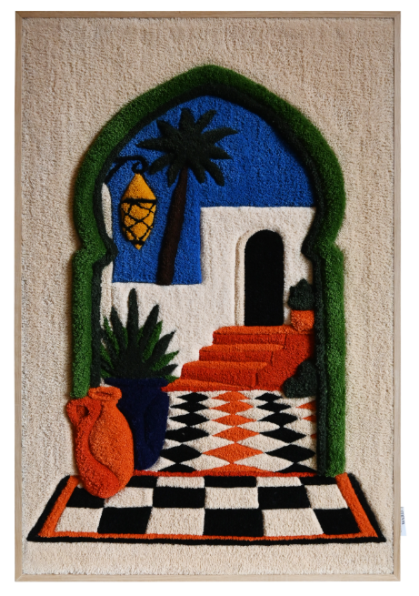 A textile portrait of a woman with a patterned scarf and closed eyes, set on a vivid blue background. The piece is displayed outdoors on a wooden easel against a brick wall with black shutters and white-framed windows