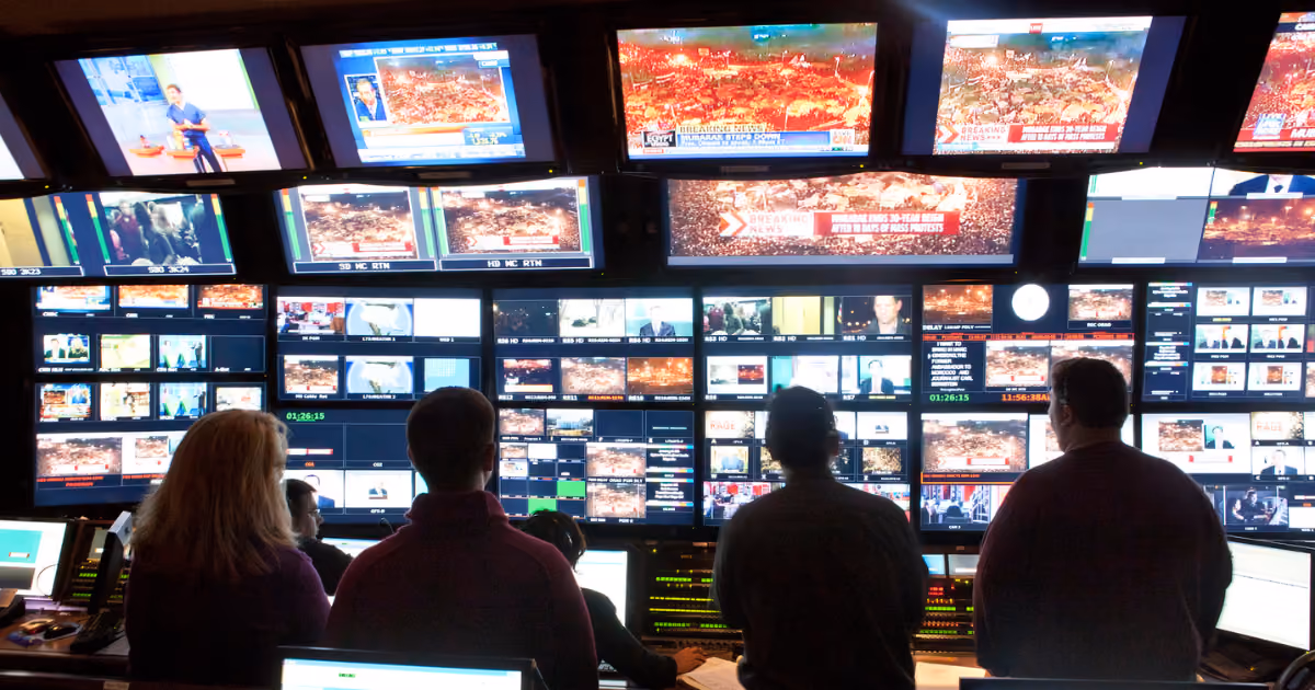 Five people monitoring multiple television screens showing breaking news and live footage in a broadcast control room.