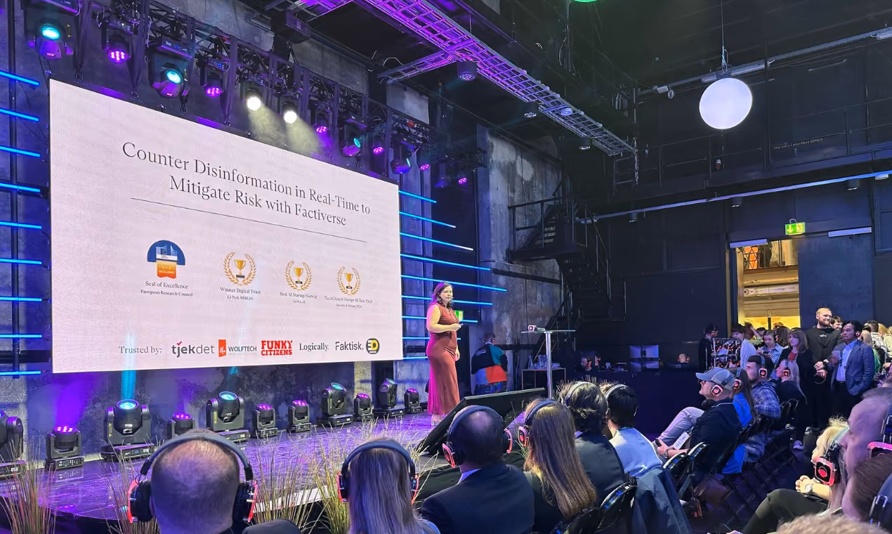 Speaker in a red dress presents on stage with a large screen behind displaying text about countering disinformation, while an audience wearing headphones listens attentively in a dimly lit venue.