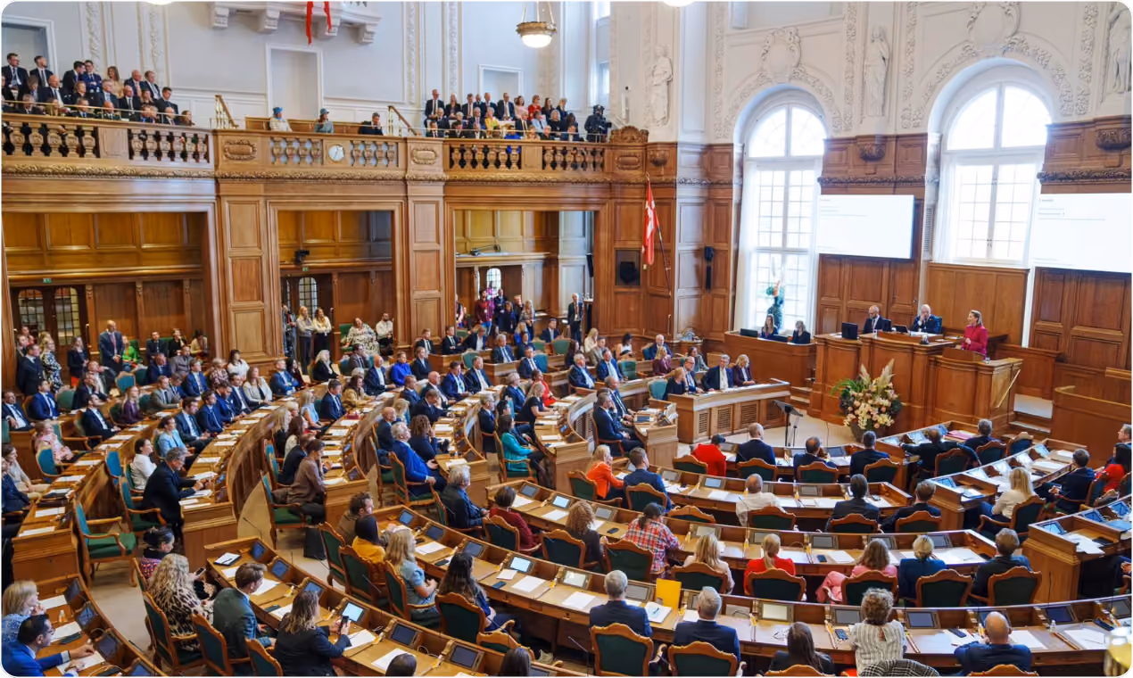 Large parliamentary chamber with members seated at curved desks, a speaker addressing from a podium, and a Danish flag displayed.
