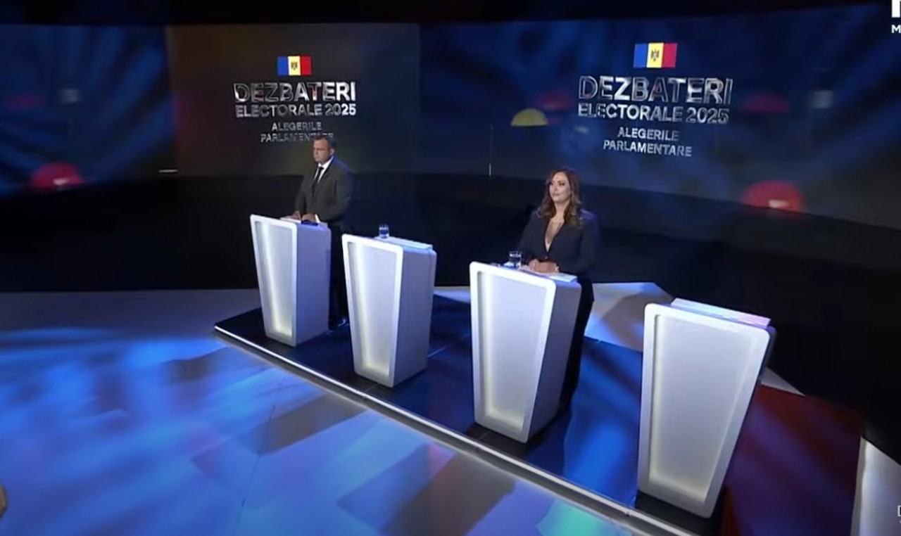 Two individuals standing behind illuminated debate podiums on a stage with screens displaying 'Dezbateri Electorale 2025 Alegerile Parlamentare' and the Moldovan flag.