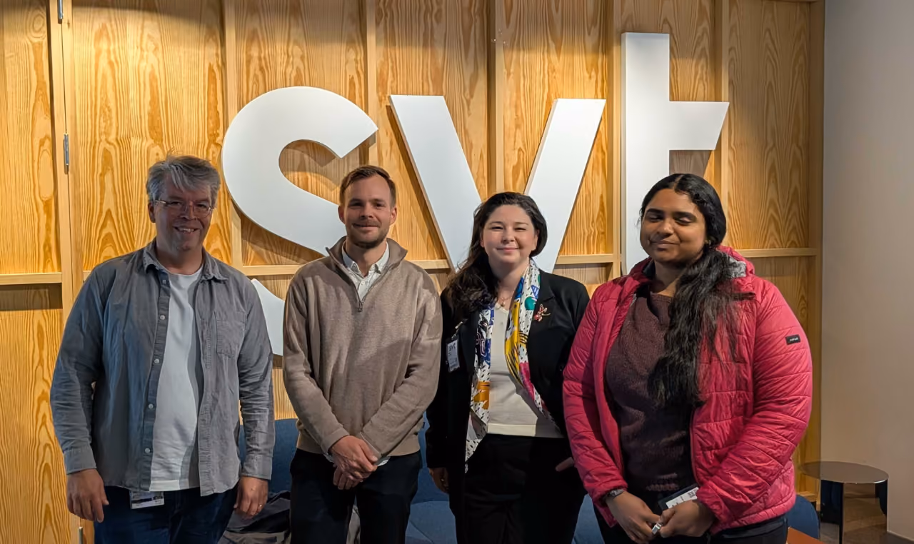 Four people standing side by side in front of a wooden wall with large white letters spelling 'svt'. They are all looking at the camera, with two men on the left and two women on the right, wearing casual and business casual clothes.