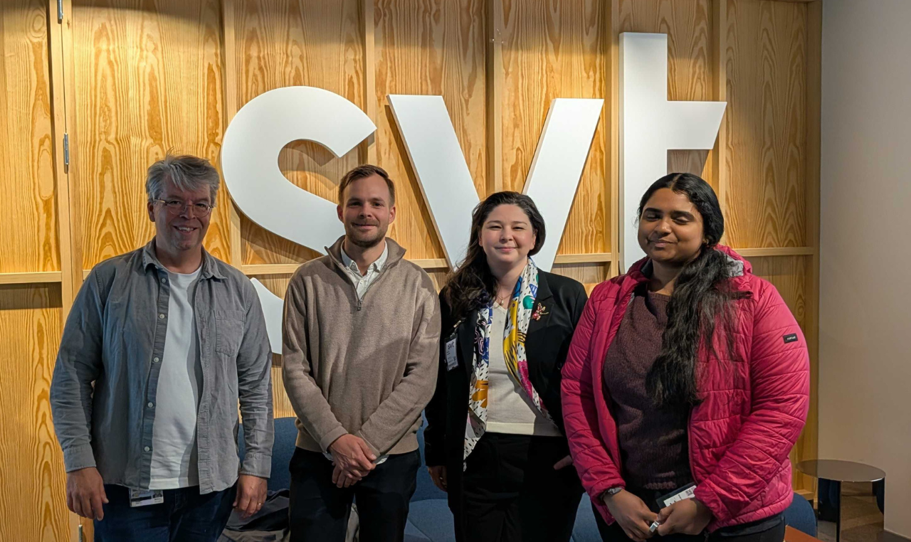 Four people standing in front of large white SVT letters on a wooden wall indoors.