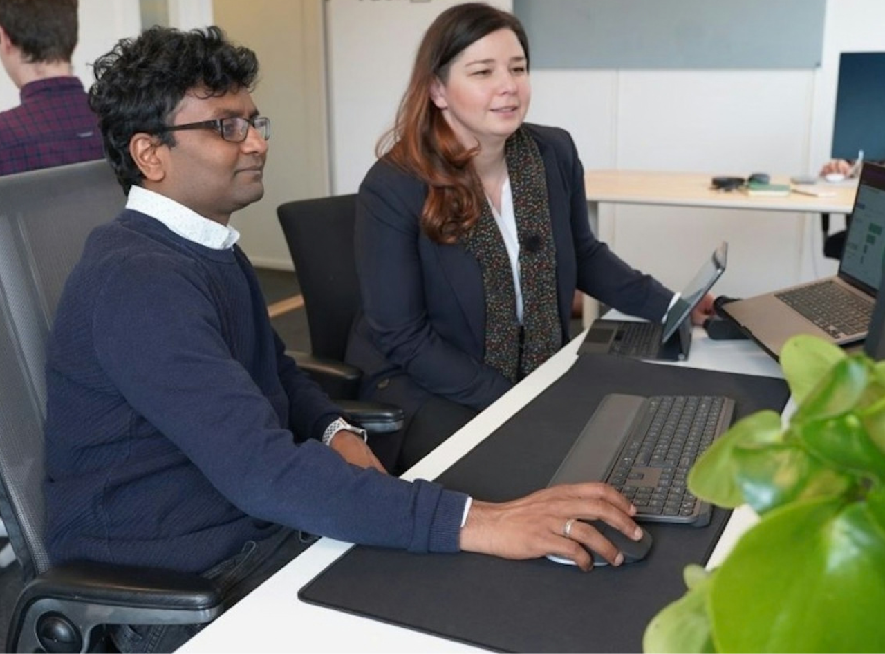A man and a woman seated at a desk collaborate while looking at a computer screen; the man is holding a mouse and the woman is holding a tablet.