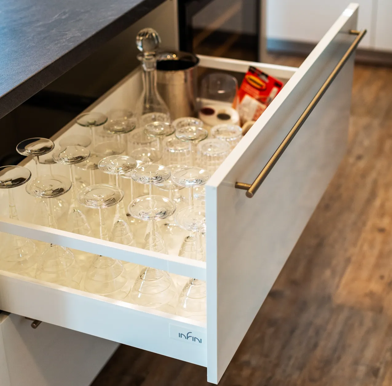Výsuvná kuchyňská zásuvka s organizérem na sklenice a karafy, plně vysunutá pod pracovní deskou, s čistým bílým provedením a kovovým madlem.