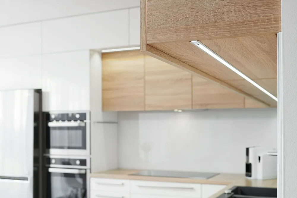 Detail horních kuchyňských skříněk s integrovaným LED osvětlením pod dřevěným korpusem, umístěných nad pracovní plochou v moderní světlé kuchyni.