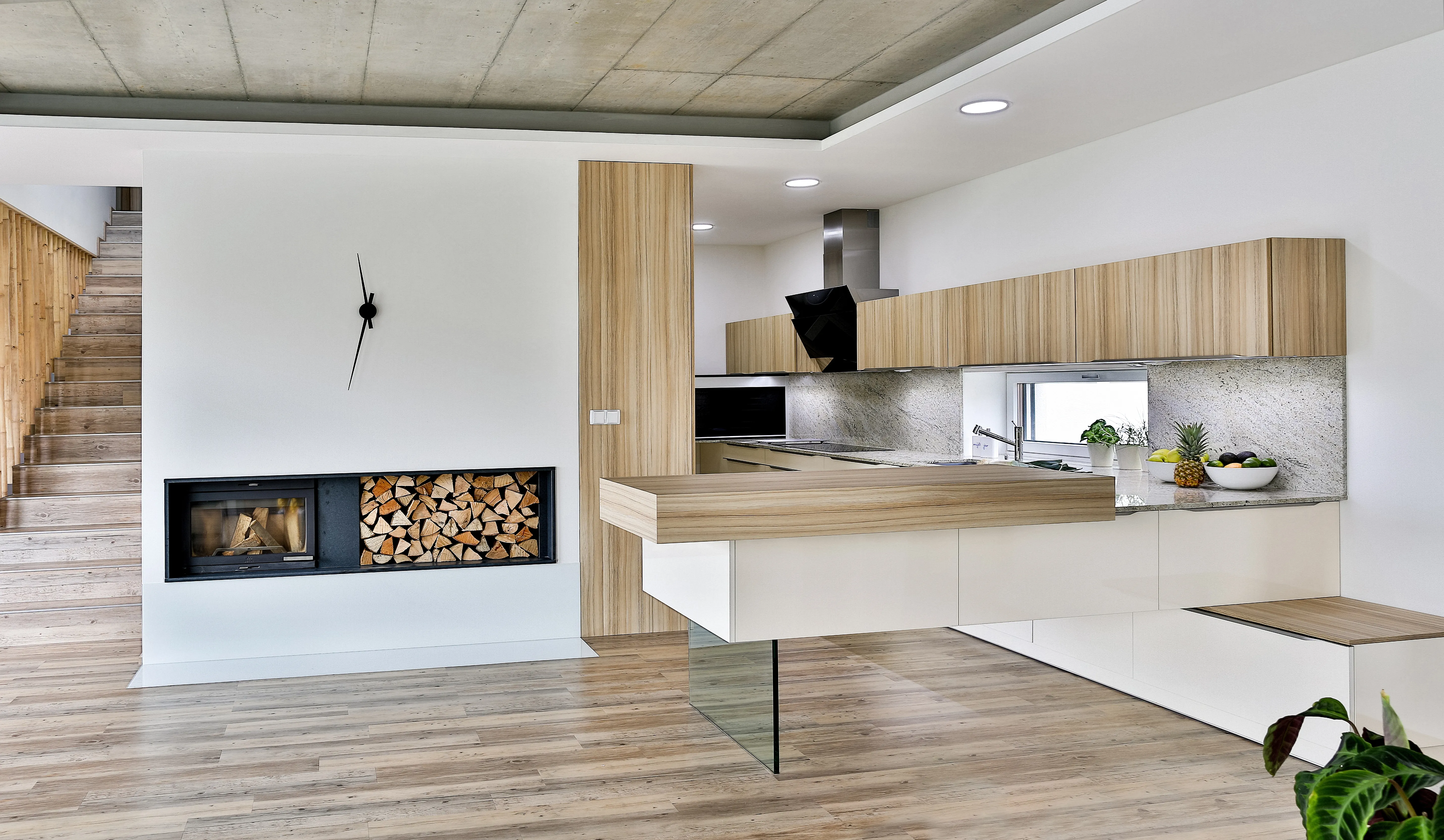 Moderní kuchyň na míru s dřevěnými skříňkami a bílými spodními zásuvkami, kamennou pracovní deskou, kuchyňským ostrůvkem s barovým přesahem, vestavěným krbem a schodištěm v otevřeném interiéru.