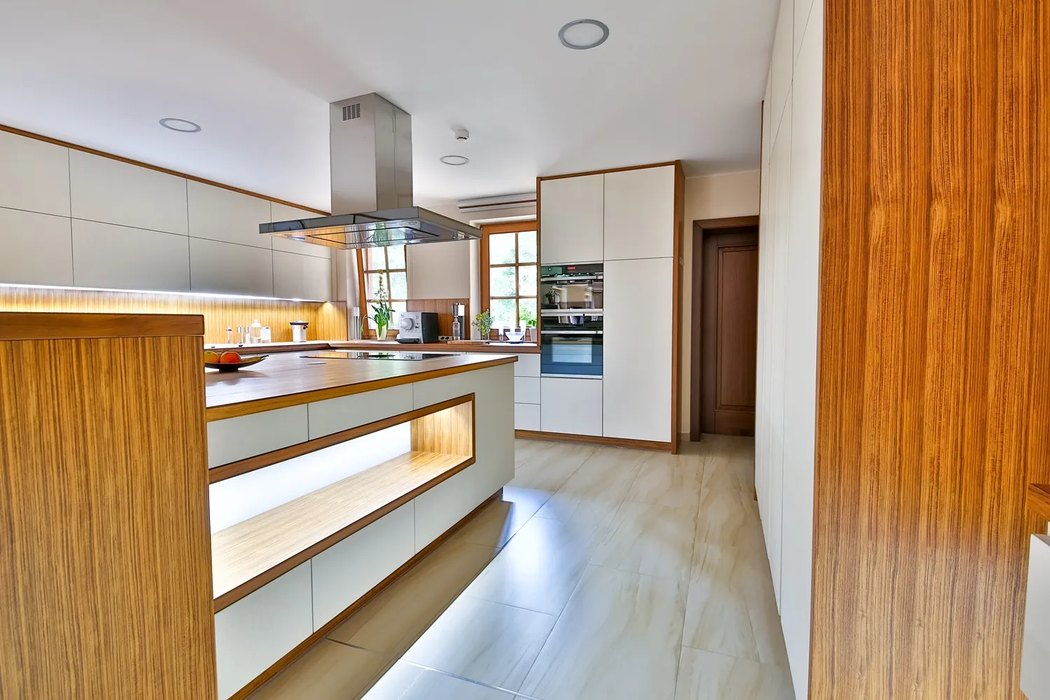 Moderní kuchyně s kombinací bílých bezúchytkových skříněk a dýhovaného dřeva, ostrůvkem s integrovaným úložným prostorem a podsvícením, dřevěnou pracovní deskou, závěsnou nerezovou digestoří a světlou dlažbou na podlaze.