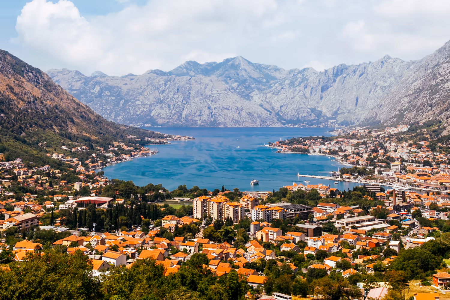 Panoramic view of a coastal town with orange rooftops, surrounded by green hills and mountains with a bay in the center.