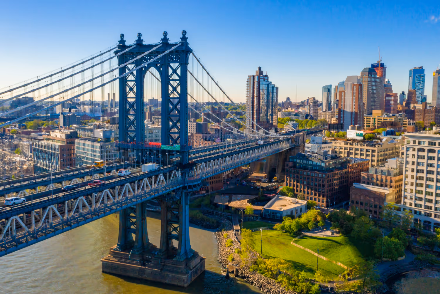 Manhattan Bridge spanning a river with city skyline and green park under clear blue sky.