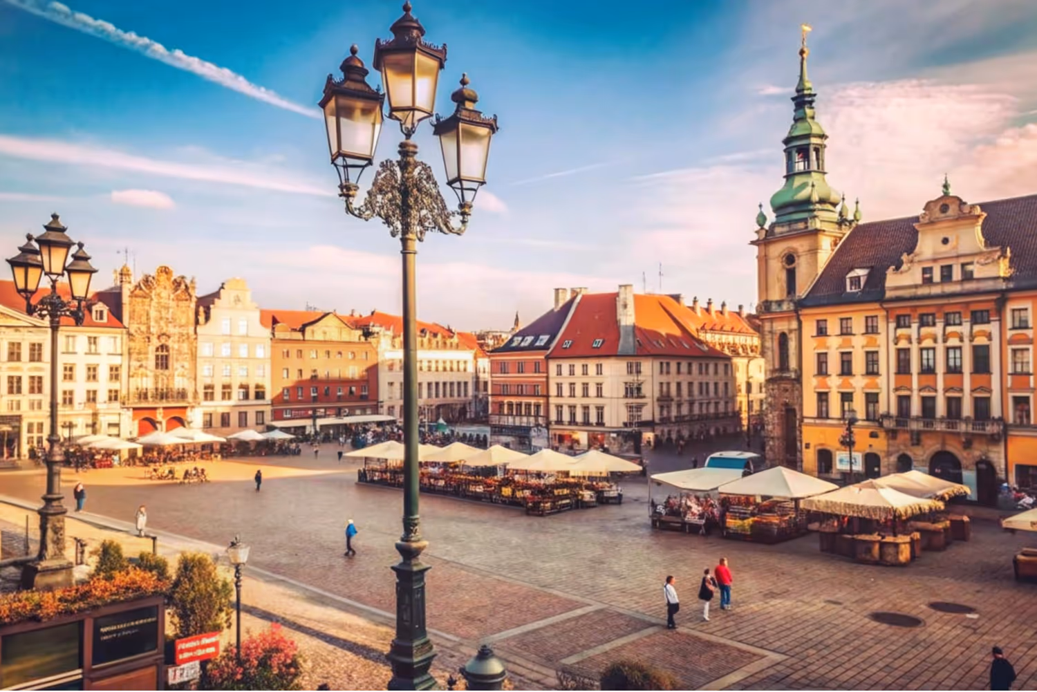 Historic European town square with ornate street lamps, market stalls under white canopies, and colorful old buildings with red roofs.