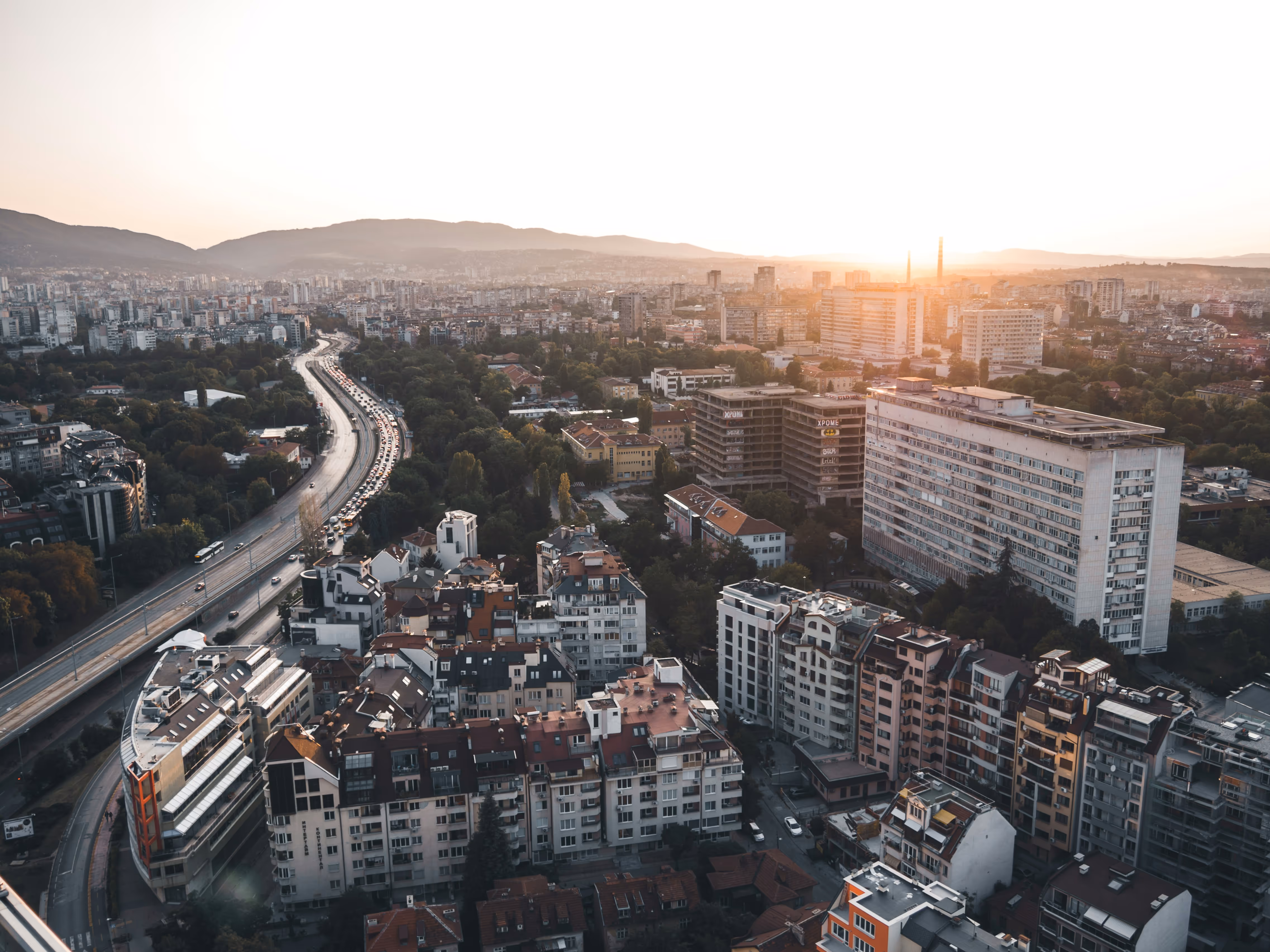 Aerial view of a cityscape with residential buildings, a curved highway with traffic, green trees, and mountains in the background at sunset.