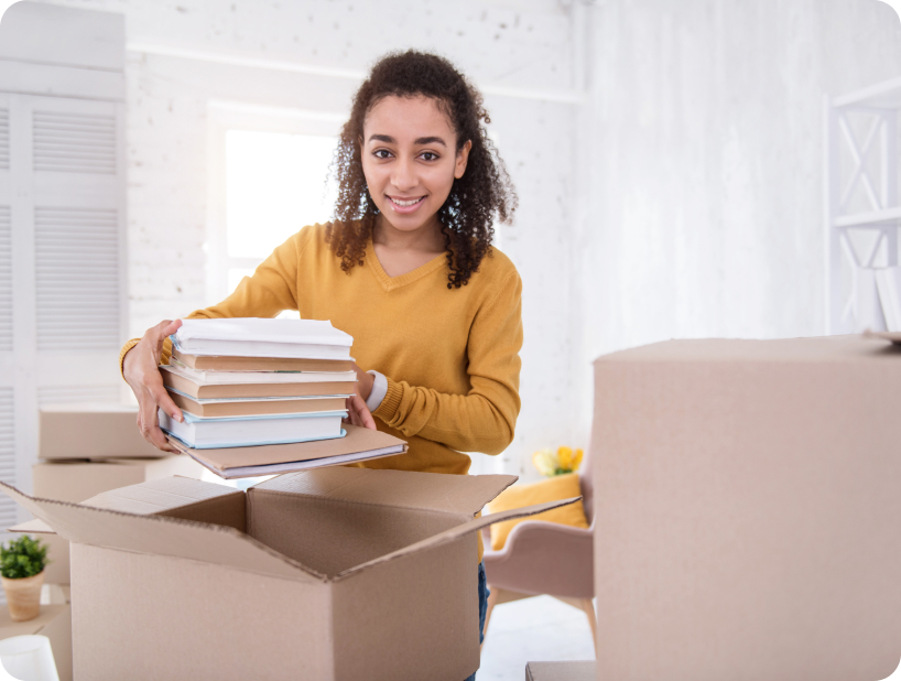 Jeune femme souriante range une pile de livres dans un carton de déménagement à l’intérieur de son nouveau logement.