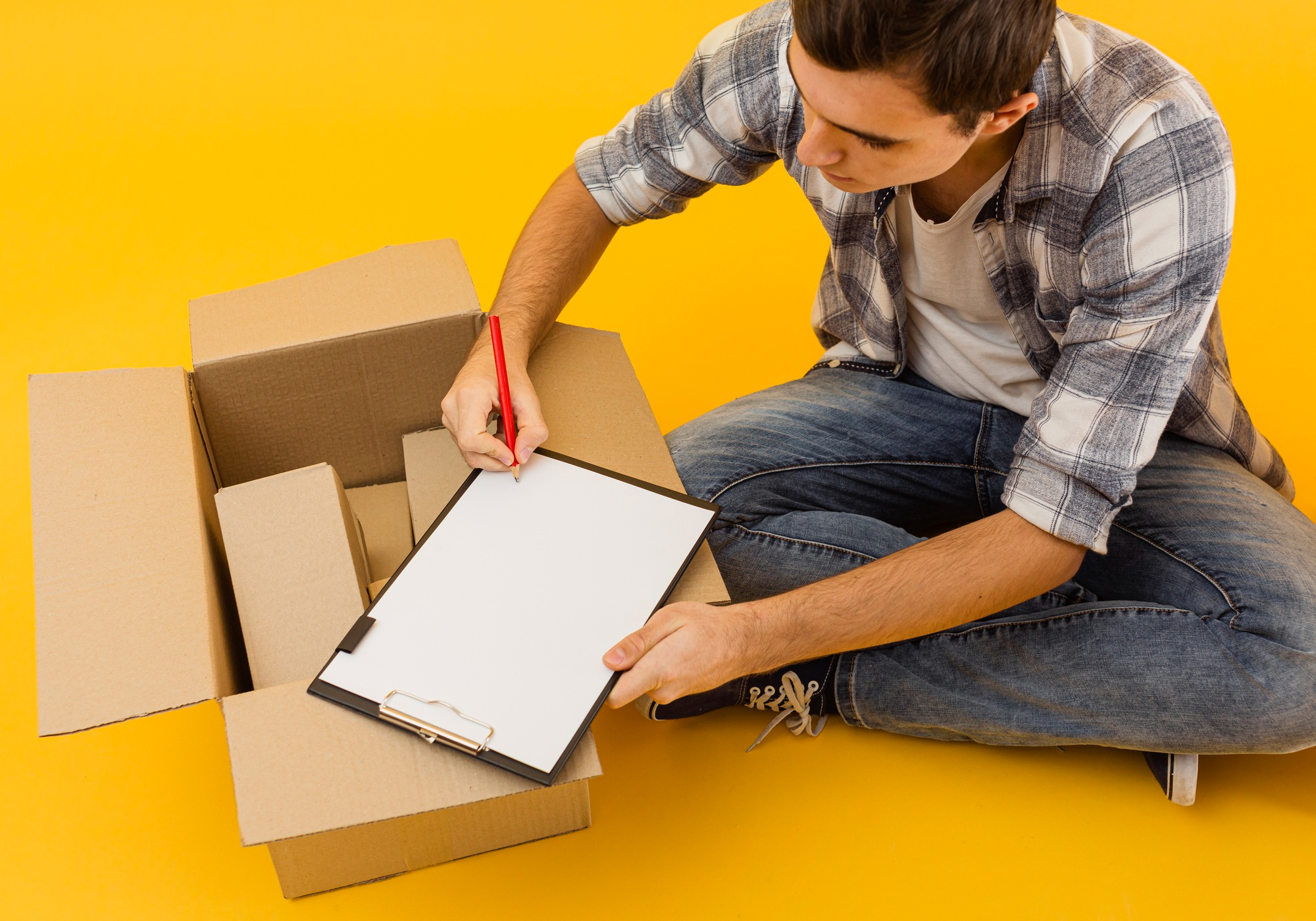 Un homme assis au sol remplit une liste sur un clipboard à côté d’une boîte ouverte pour préparer son déménagement efficacement.