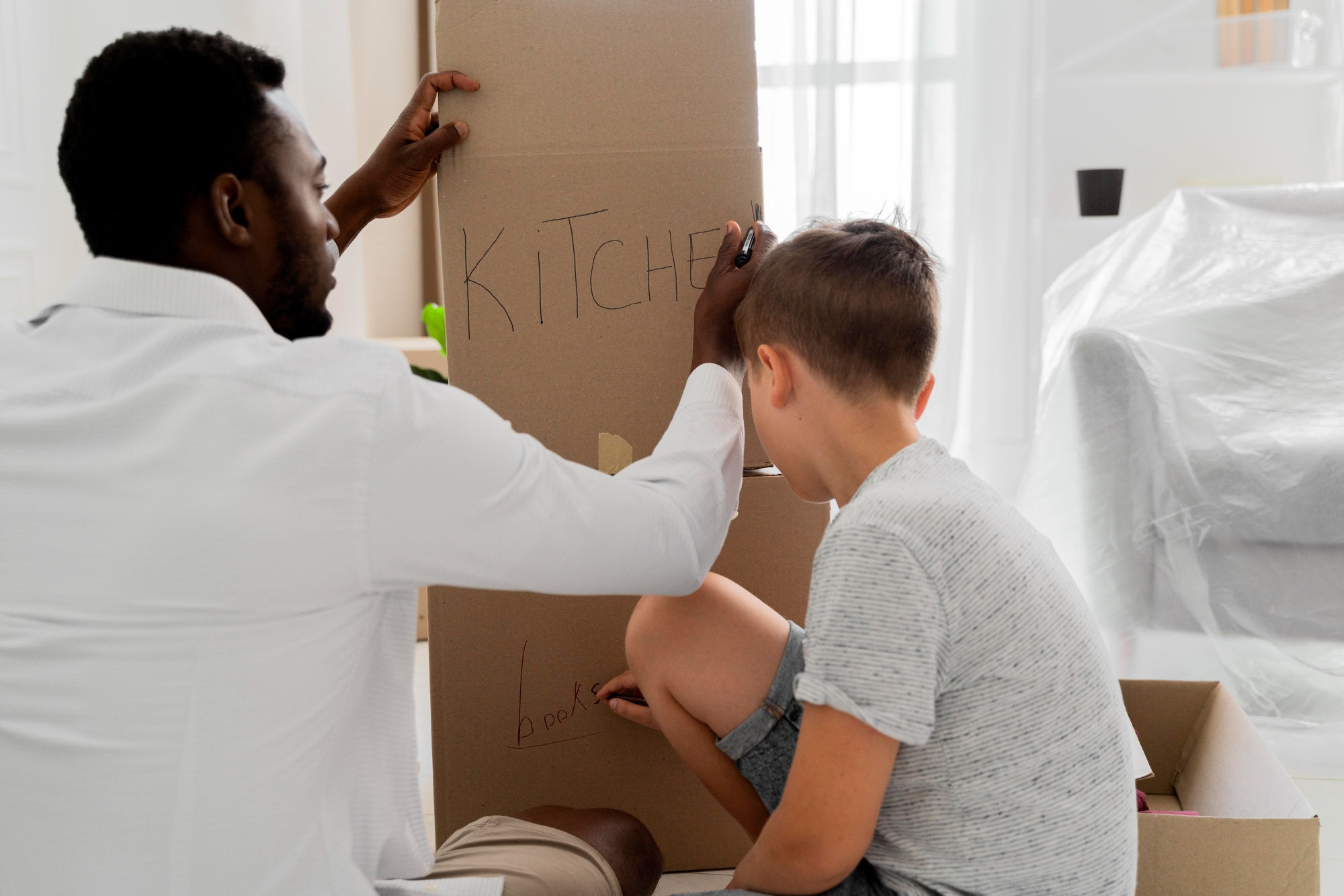 Un adulte aide un enfant à écrire « Kitchen » sur une grande boîte en carton afin de préparer son déménagement avec sa famille.