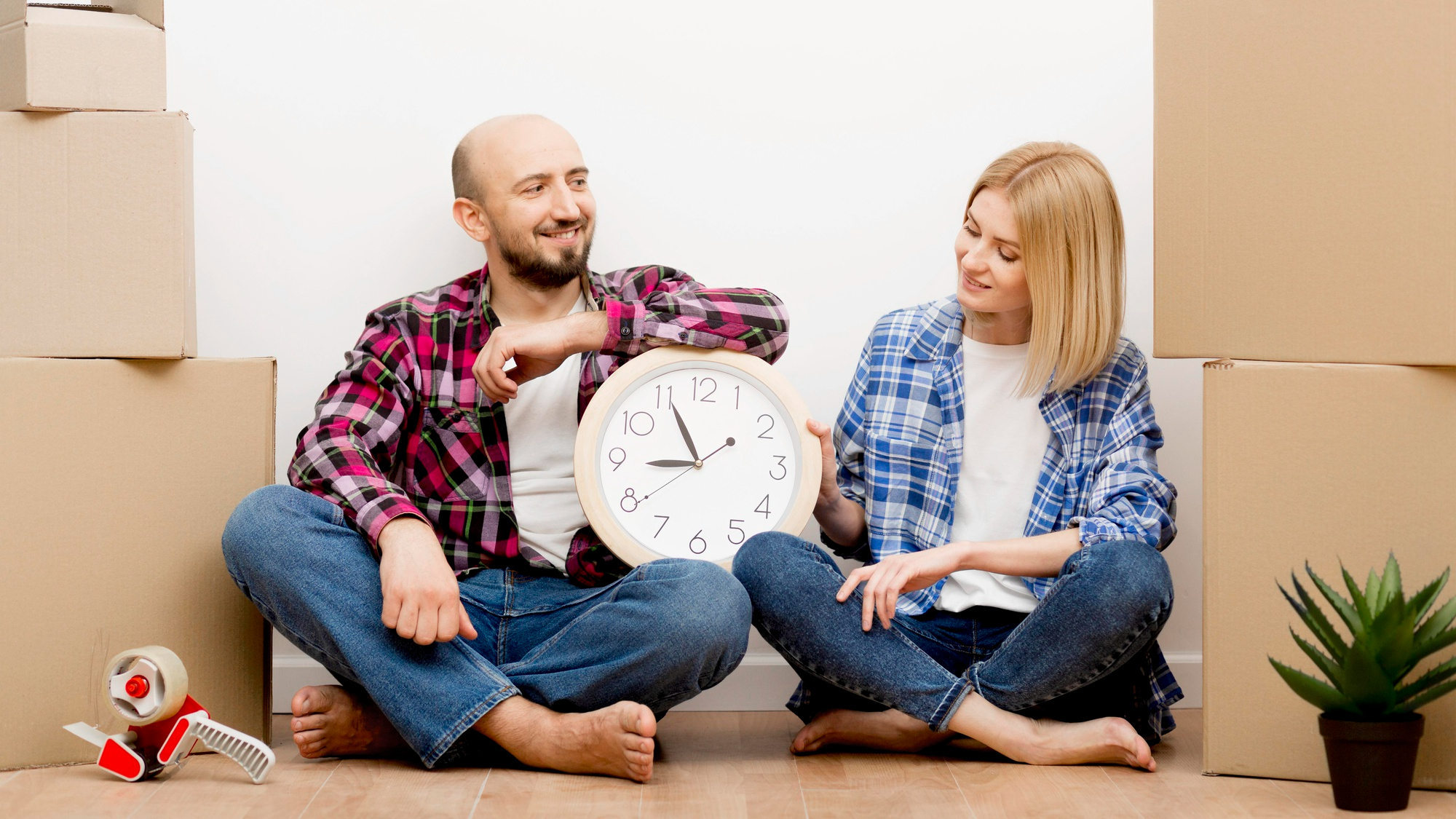 Couple assis au sol entre des boîtes de déménagement, souriant en regardant une horloge, illustrant la planification et le bon timing avant un déménagement.