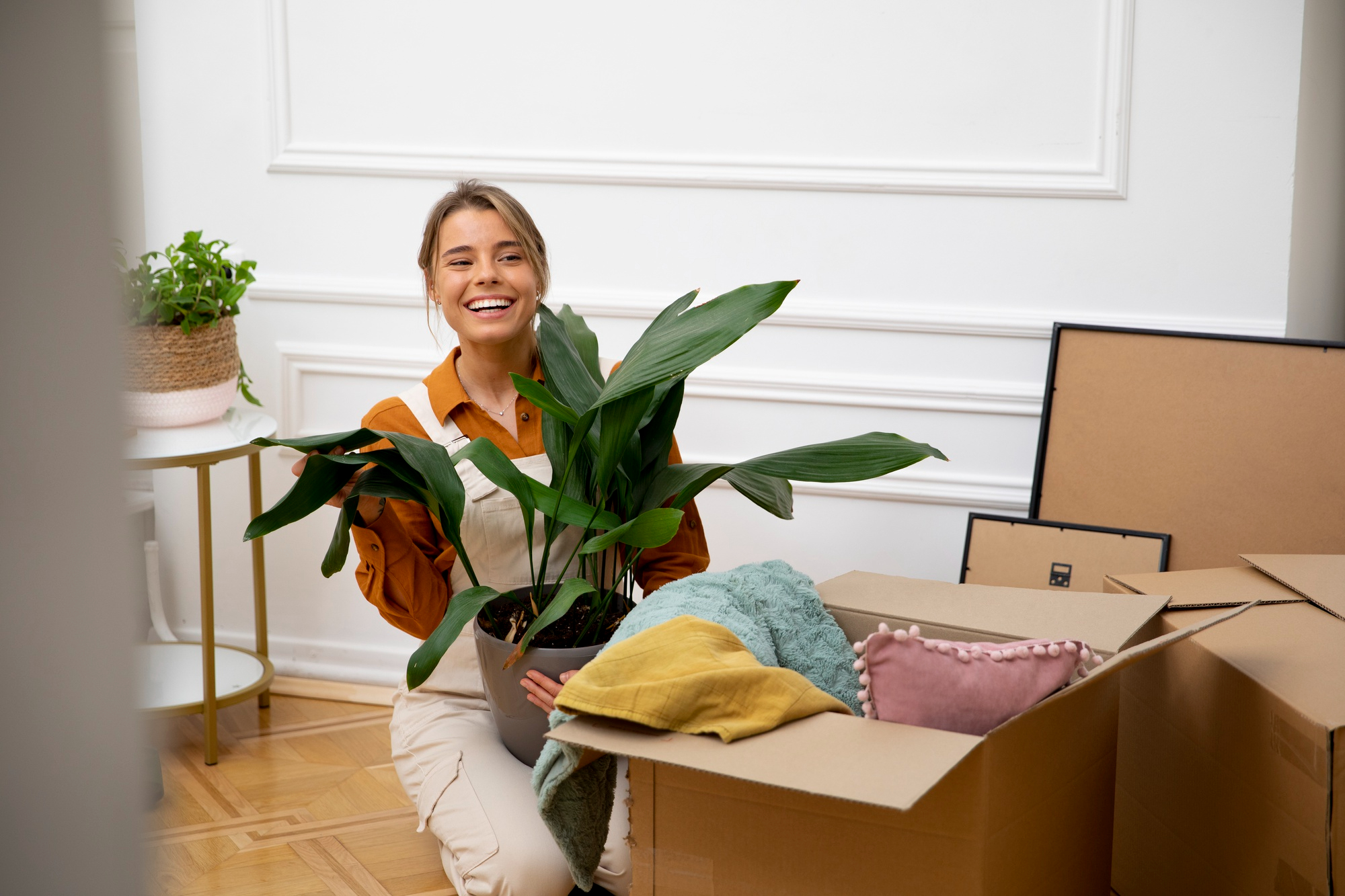 Femme souriante tenant une plante verte devant des boîtes ouvertes, illustrant un déménagement réussi et une installation positive dans un nouveau logement.