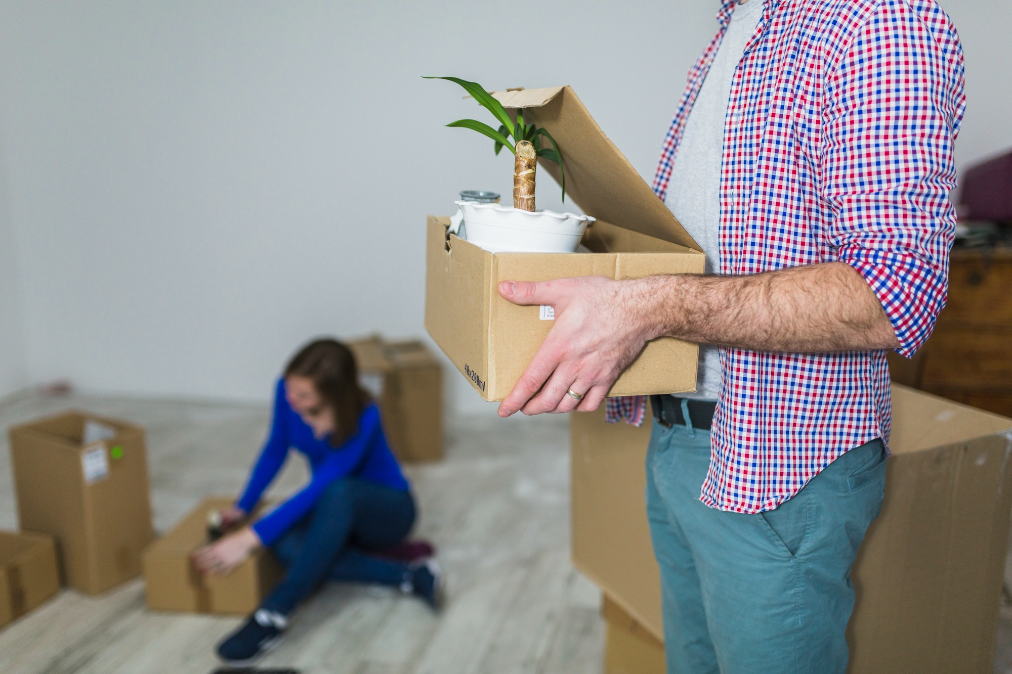 Homme tenant une boîte avec une plante pendant qu’une femme organise des cartons à l’arrière, illustrant un déménagement résidentiel en cours