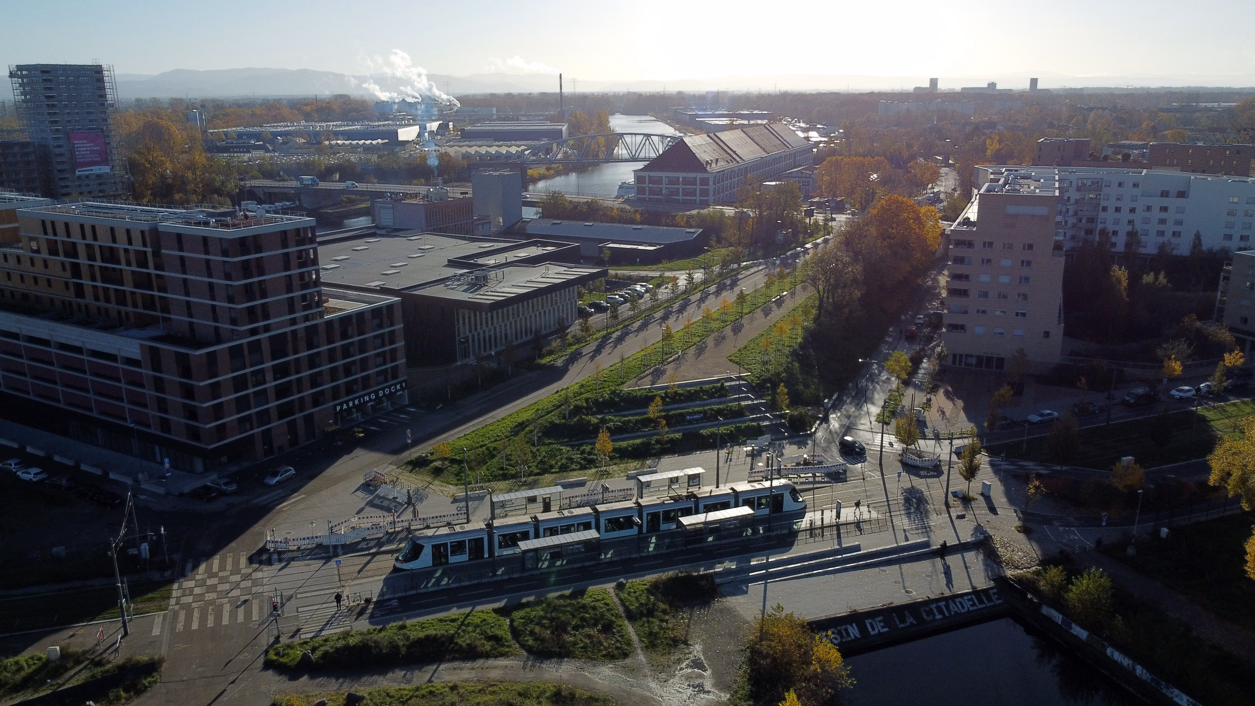 Vue aérienne d'un quartier urbain traversé par une ligne de tramway, avec des bâtiments modernes, des arbres aux couleurs automnales et un canal au premier plan