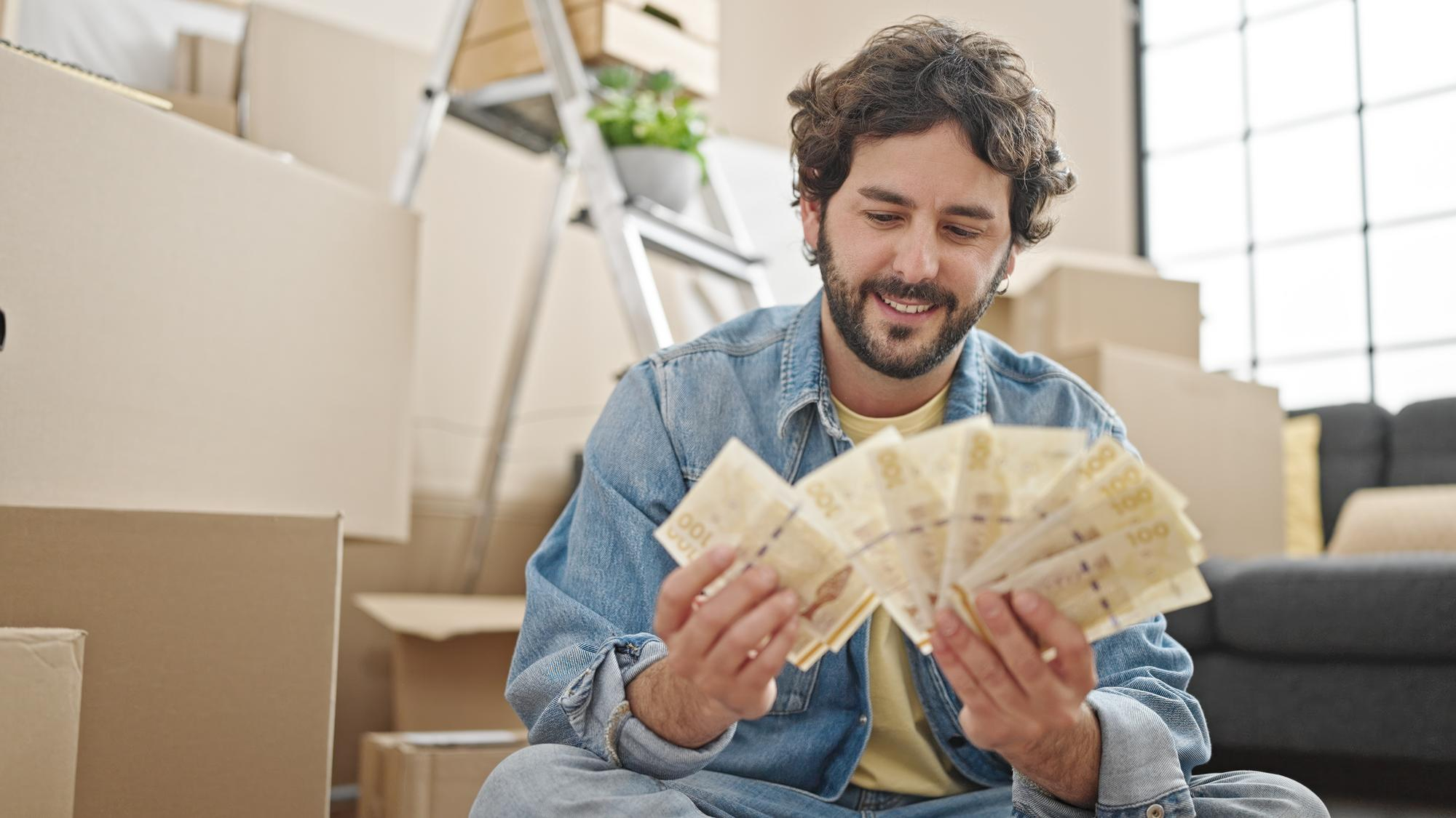 Homme souriant, comptant des billets de banque, assis au sol parmi des boîtes de déménagement, évaluant le coût de son projet de déménagement.