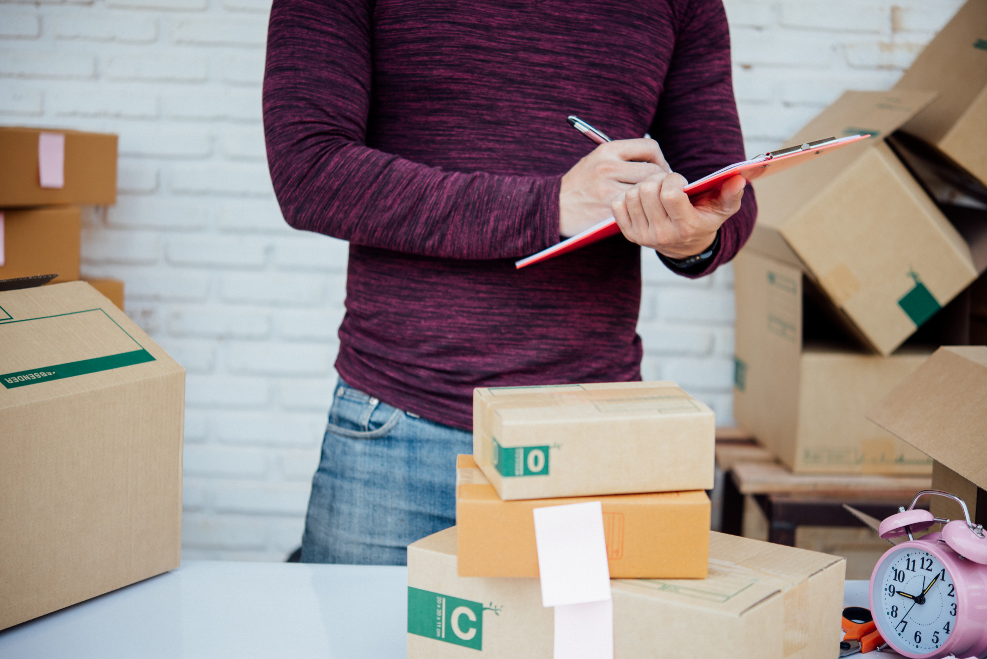 Personne prenant des notes sur un bloc devant des boîtes de carton empilées, préparant l'inventaire de ses biens avant un déménagement.