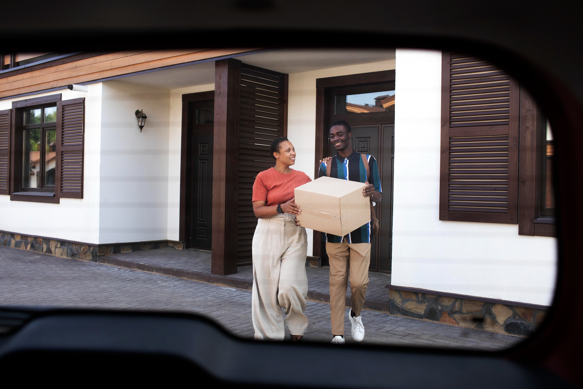 Un couple souriant transportant ensemble une boîte de déménagement en carton devant une maison aux volets fermés, encadré par la vitre arrière d'une voiture.