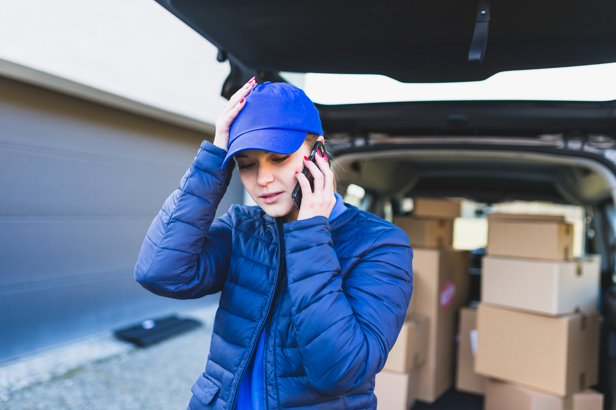 Déménageuse en veste bleue au téléphone, devant un coffre de voiture rempli de boîtes, lors d'un déménagement d'urgence.