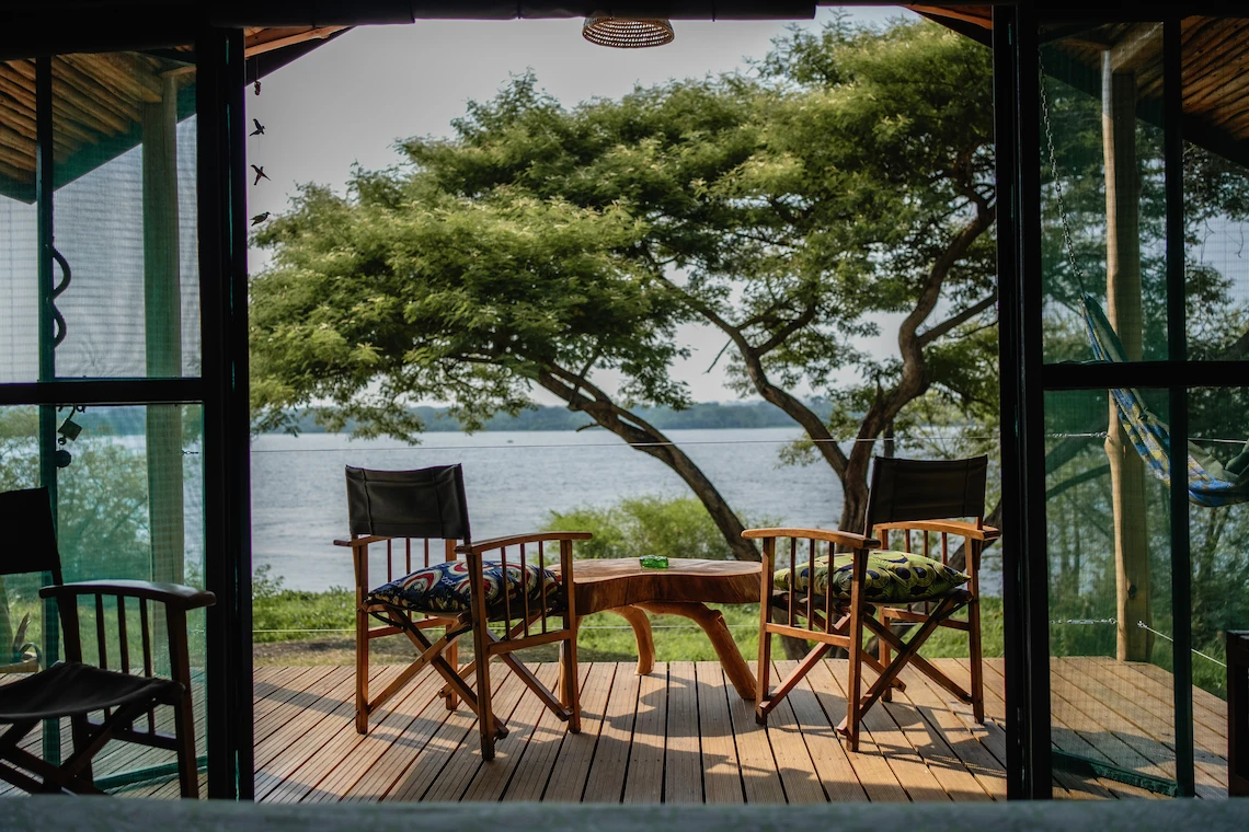 Two chairs and a natural wood live-edge coffee table on the deck of a glass-fronted balcony, with a hammock and views overlooking the River Nile.