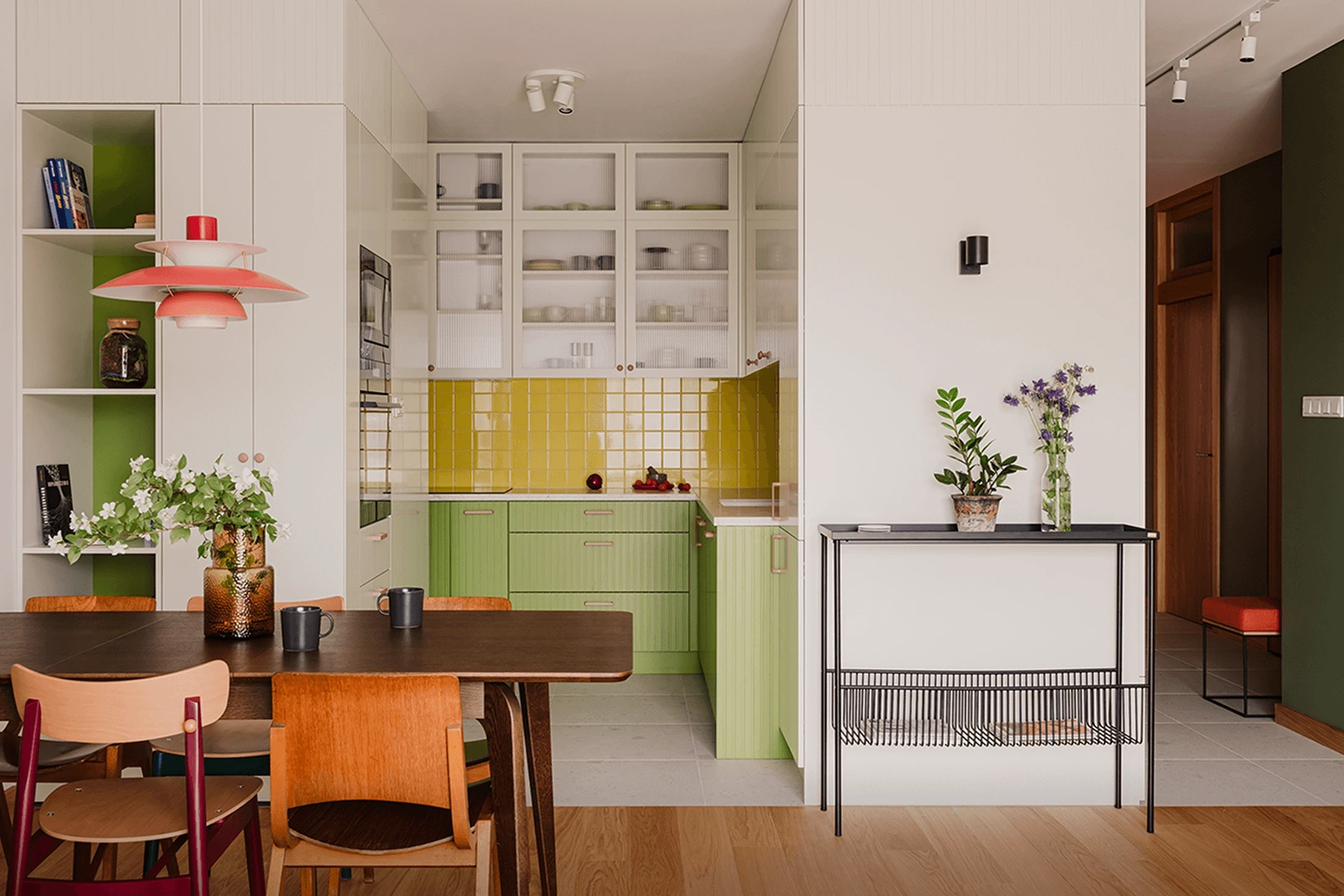 Jasna kuchnia z zielonymi szafkami i żółtymi kafelkami, ciemny stół z różnorodnymi drewnianymi krzesłami i kwiaty na stole oraz na metalowej półce przy ścianie.