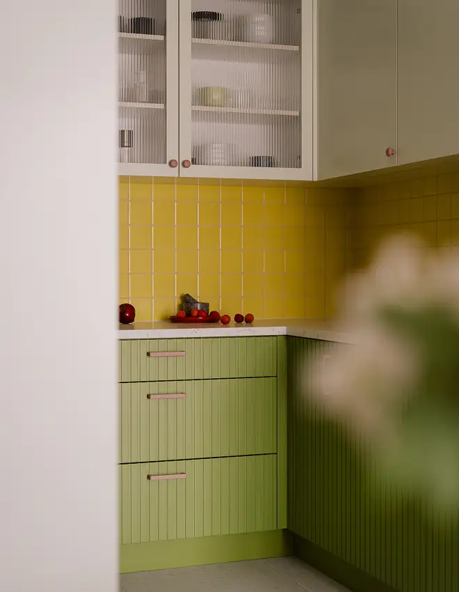Widok na zielone szafki kuchenne z żółtymi kafelkami na ścianie oraz przezroczystymi szafkami górnymi z naczyniami w środku.