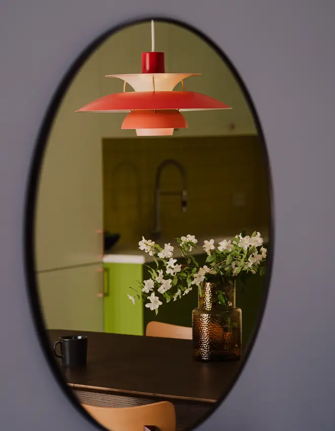 Widok przez owalne lustro na kwiaty w wazonie na stole, czerwony zwisający kinkiet i kuchnię w tle.