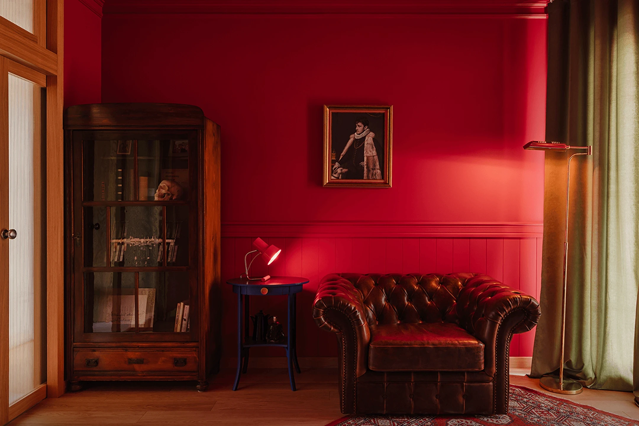 Przytulny pokój z bordowymi ścianami, skórzanym brązowym fotelem, drewnianą witryną, niebieskim stolikiem z czerwoną lampką oraz obrazem na ścianie.