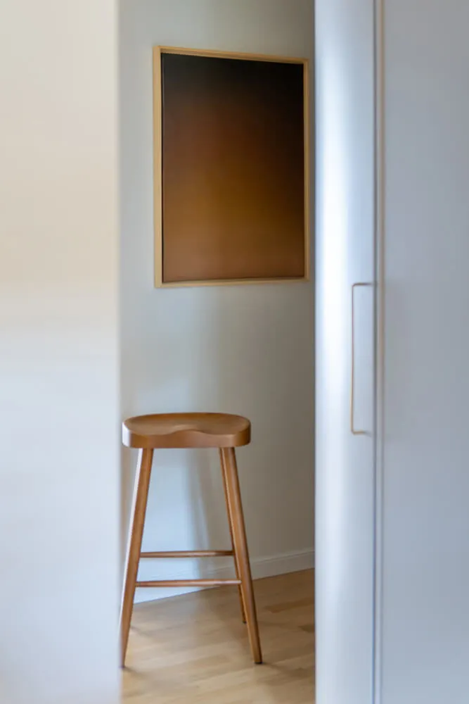 Drewniany stołek z oparciem na drewnianej podłodze obok białej ściany i ciemnego obrazu w jasnej, minimalistycznej przestrzeni.