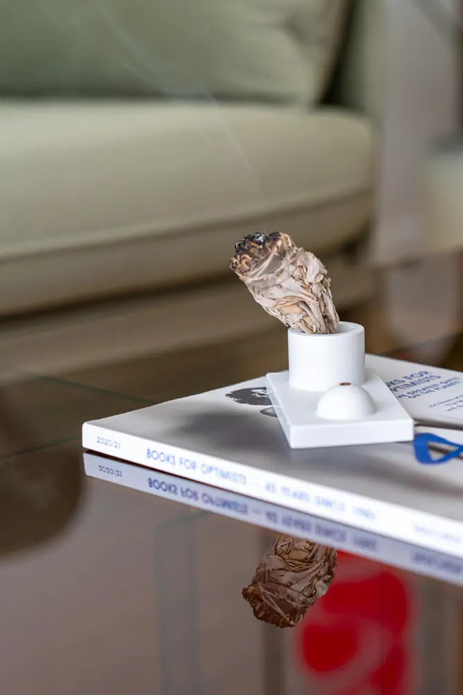 Płonąca biała szałwia w ceramicznym kadzidełku na stosie książek na szklanym stoliku, w tle rozmyta sofa.