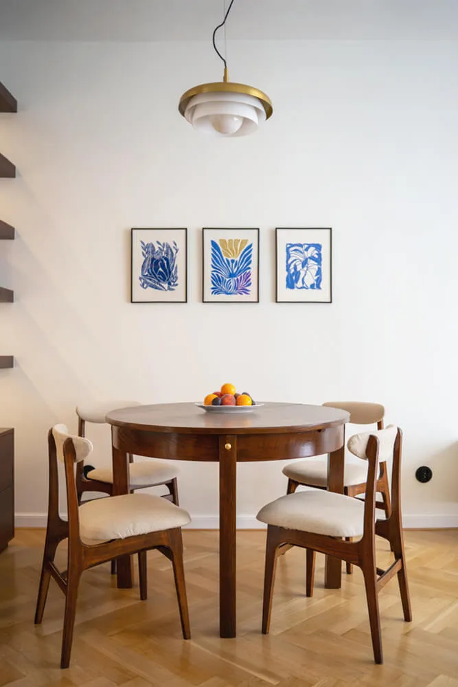 Jadalnia z drewnianym okrągłym stołem, czterema tapicerowanymi krzesłami, owocami na talerzu i trzema grafikami na ścianie.
