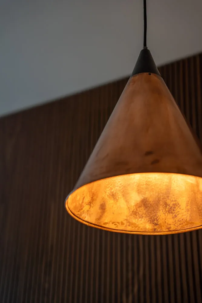 Wisząca metalowa lampa w kształcie stożka z ciepłym, pomarańczowym światłem, na tle ciemnej, drewnianej ściany.